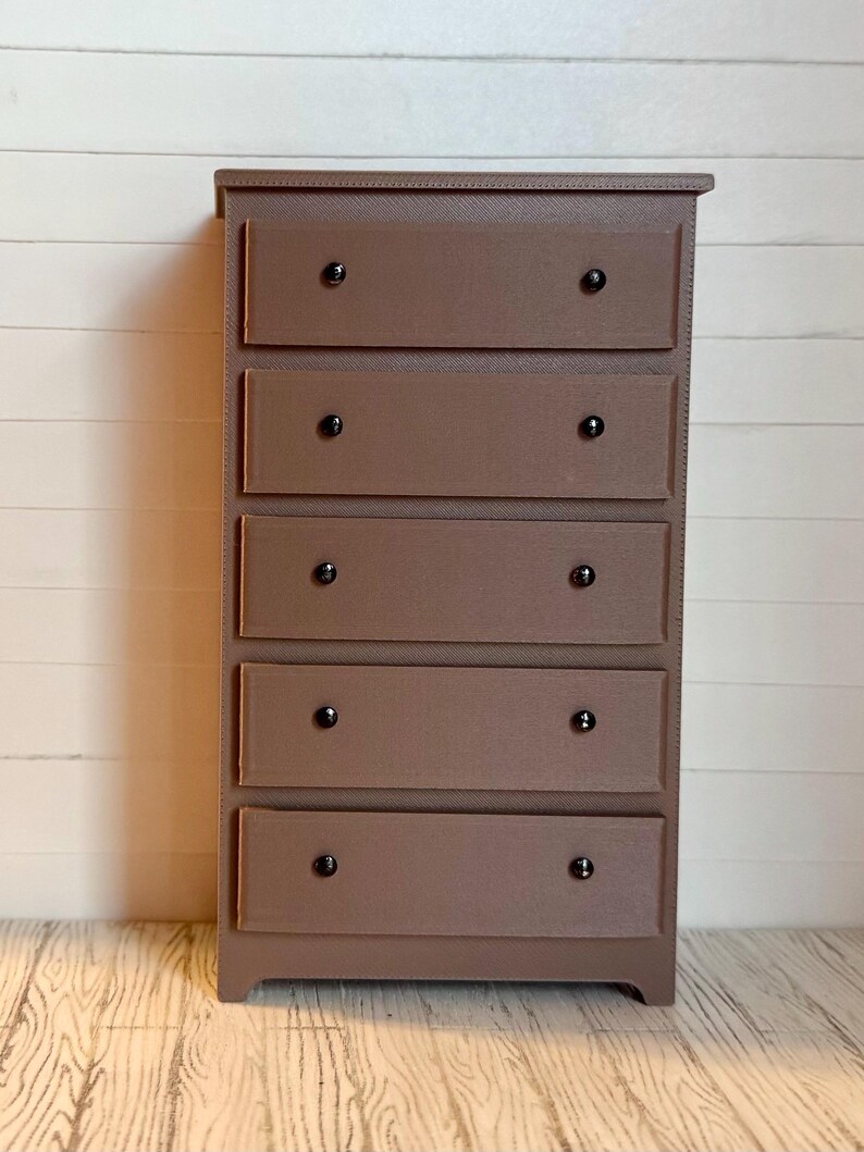 May include: A brown wooden chest of drawers with five drawers and black knobs. The chest of drawers is standing on a light wood floor.