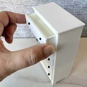 May include: White wooden chest of drawers with five drawers and black knobs. The top drawer is open.