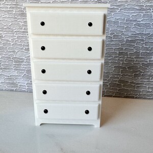May include: White wooden chest of drawers with five drawers and black knobs. The chest of drawers has a simple design and is made of solid wood.
