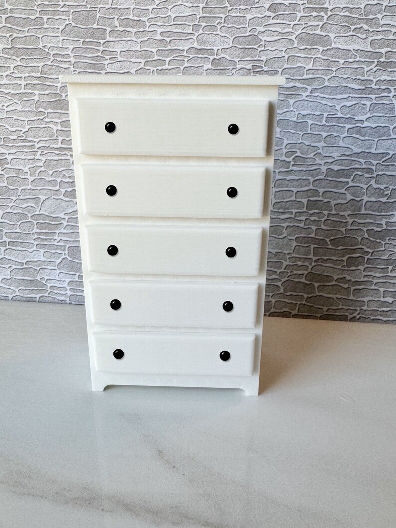 May include: White wooden chest of drawers with five drawers and black knobs. The chest of drawers has a simple design and is made of solid wood.