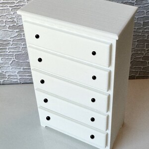 May include: White chest of drawers with five drawers and black knobs. The chest of drawers is made of wood and has a simple design.
