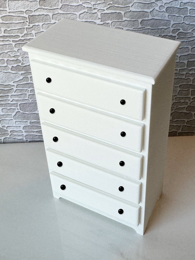 May include: White chest of drawers with five drawers and black knobs. The chest of drawers is made of wood and has a simple design.