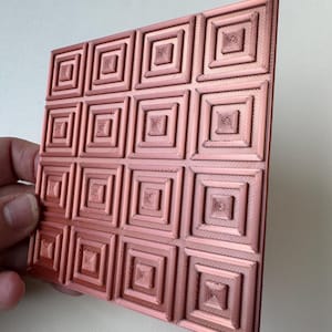 May include: A copper-colored, square decorative panel featuring a repeating pattern of nested squares. The raised design creates a textured surface. The panel is held at an angle, showcasing the geometric pattern.