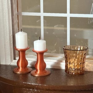 May include: Two white pillar candles sit atop copper-colored candle holders, next to a textured amber glass. The items are arranged on a dark brown, semi-circular table, with a window in the background.