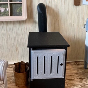 May include: A miniature black stove with a silver door and vertical vents. A small wooden bucket holds several pieces of firewood. The stove has a black chimney and sits in a miniature room setting.