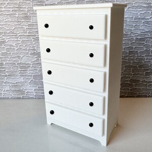 May include: White chest of drawers with five drawers and black knobs. The chest of drawers is made of wood and has a simple design.