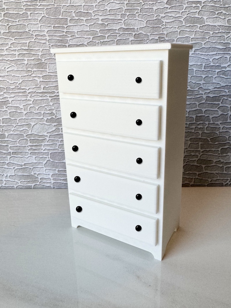 May include: White chest of drawers with five drawers and black knobs. The chest of drawers is made of wood and has a simple design.