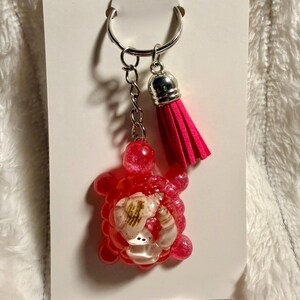 May include: A red resin turtle keychain with seashells embedded inside. The keychain has a silver keyring, chain, and a bright pink tassel. The turtle is attached to a white card.