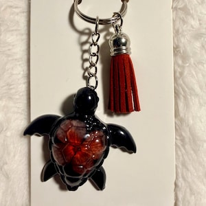 May include: A black turtle keychain with a red and orange floral design on the shell. The keychain includes a silver chain, a red tassel, and a silver key ring. The turtle has outstretched flippers.