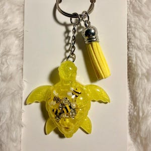 May include: A yellow resin turtle keychain with a silver keyring and chain. The turtle has a bee and jewel embellishment. A yellow tassel hangs from the chain. The keychain is on a white card.