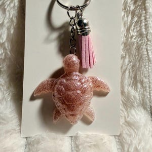 May include: A pink glitter turtle keychain with a silver keyring, chain, and tassel. The turtle has outstretched flippers and a textured shell. The tassel is a light pink color.