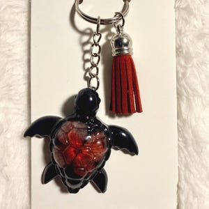 May include: A keychain featuring a black sea turtle charm with a red pressed flower embedded in its shell. The charm is attached to a silver-toned metal split ring and chain, which also holds a small, rust-colored suede leather tassel.
