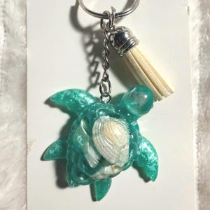 May include: A teal turtle keychain with a seashell center and a silver keyring. The turtle is made of resin and has a tassel. The keychain is a decorative accessory.