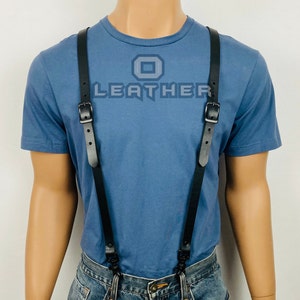 Puede incluir: Tirantes de cuero negro con hebillas y clips metálicos. Los tirantes se llevan sobre una camiseta azul con la palabra "LEATHER" impresa.