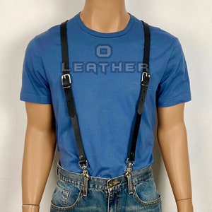Könnte beinhalten: Schwarze Lederhosenträger mit silbernen Schnallen und Clips. Die Hosenträger werden über einem blauen T-Shirt und einer blauen Jeans getragen.