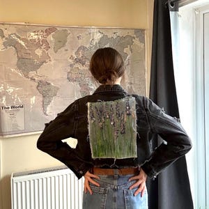 Può includere: Giacca di jeans nera con un disegno floreale dipinto sul retro. L'opera d'arte presenta fiori viola ed erba verde. La giacca ha una vestibilità classica con chiusura a bottoni e dettagli strappati.