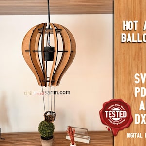 Könnte beinhalten: Eine Holzlampe in Form eines Heißluftballons mit einem schwarzen Kabel, das von der Decke hängt. Die Lampe ist aus lasergeschnittenem Holz und hat einen kleinen Korb, der an der Unterseite hängt. Die Lampe steht auf einer Holzoberfläche mit einer kleinen Pflanze in einem Topf daneben. Der Text "HOT AIR BALLOON" ist in Weiß auf braunem Hintergrund geschrieben. Der Text "SVG PDF AI DXF DIGITAL FILES" ist in Weiß auf braunem Hintergrund geschrieben.