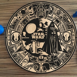 May include: A round wooden coaster with a black and white design featuring characters from Star Wars. The design includes Darth Vader, Yoda, and the Millennium Falcon. The text "STAR WARS" is printed in the center of the coaster.