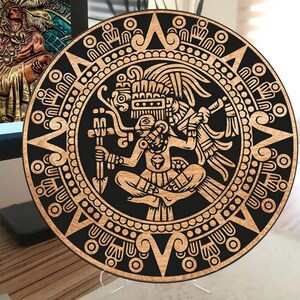 Puede incluir: Un colgante de pared circular de madera con un diseño en negro y marrón. El diseño presenta una figura estilizada con alas y un borde circular con patrones repetidos.