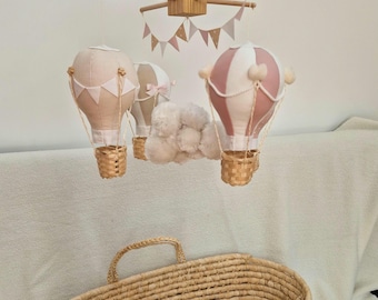 May include: A pink and white striped hot air balloon mobile with a cloud and a banner of flags hanging from a wooden frame. The mobile is hanging above a woven basket.