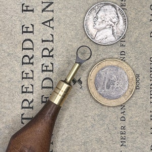May include: A wooden tool with a brass tip and a loop at the end, a silver US half dollar coin with the profile of a man, and a silver Euro coin with the number "1" on it. The coins are on a piece of paper with text in Dutch.