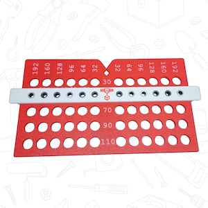 Può includere: Uno strumento rosso con più fori e misure. Lo strumento ha una barra bianca con fori e numeri aggiuntivi. I numeri includono 30, 32, 64, 70, 90, 96, 110, 128, 160 e 192. Lo strumento è progettato per misurare e contrassegnare.