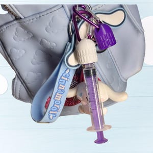 Pu&ograve; includere: Una collezione di portachiavi e ciondoli, tra cui una siringa viola, un tappo di bottiglia bianco e viola e un cinturino blu con la scritta "Cinnamoroll". Gli articoli sono attaccati a una borsa azzurra con dettagli a forma di nuvola.