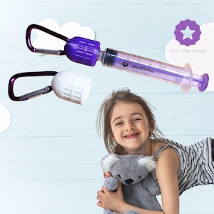Pu&ograve; includere: Una siringa per medicinali viola e trasparente con un tappo bianco e un moschettone viola. Sulla siringa &egrave; stampata la parola "medicina". L'immagine include anche una bambina che tiene in braccio un koala di peluche grigio.