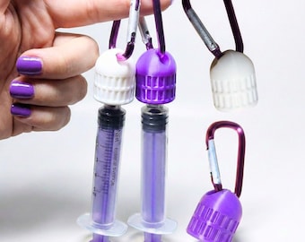 May include: Purple and white syringe-shaped containers with carabiner clips. The containers have a clear, graduated barrel and a purple plunger. The caps are either white or purple. The image is on a white background.