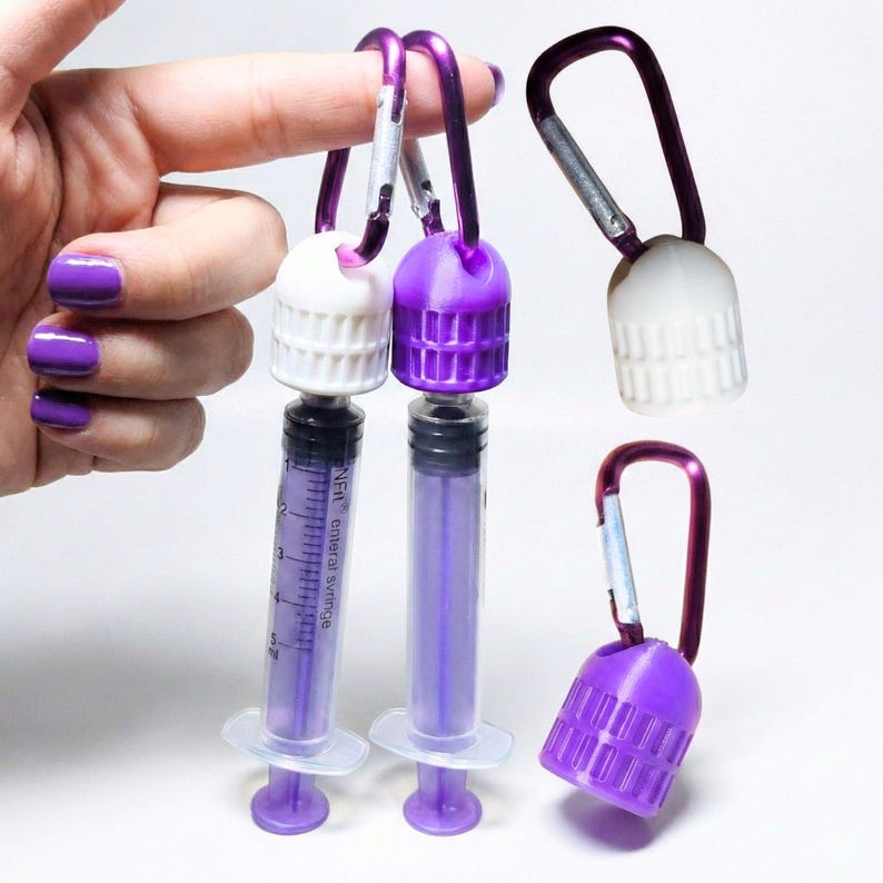 Pu&ograve; includere: Contenitori a forma di siringa viola e bianchi con moschettoni. I contenitori hanno un corpo trasparente graduato e uno stantuffo viola. I tappi sono bianchi o viola. L'immagine &egrave; su sfondo bianco.