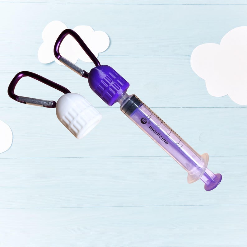 Pu&ograve; includere: Una siringa viola e trasparente con la scritta "medicina" stampata sopra, attaccata a un moschettone viola. Viene mostrato anche un tappo bianco con un moschettone viola. Gli oggetti sono posizionati su uno sfondo azzurro con ritagli di nuvole bianche.