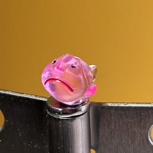 May include: A small, translucent pink glass figurine shaped like a fish with a distinct downturned mouth, perched atop a brushed silver metal door hinge that features two circular screw holes on each side of the central cylindrical barrel.
