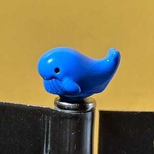 May include: A small, bright blue whale-shaped figurine with a smooth, glossy finish and a tiny black dot for an eye. The whale is positioned on top of a dark, metallic hinge component that features a circular cutout on each side of the metal plate.