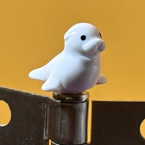 May include: A small white seal figurine with black eyes and a smooth finish, mounted on top of a metallic hinge pin. The hinge features two rectangular metal plates, each with a circular hole, connected by a central cylindrical barrel component.