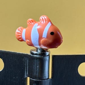 May include: A small, orange and white clownfish-shaped decorative topper attached to the top of a metallic hinge. The fish features a smooth, glossy finish with distinct white vertical stripes and a single black dot representing the eye.