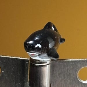 May include: A small, glossy black and white orca figurine with a smiling expression, featuring a dorsal fin and side fins, mounted atop a cylindrical silver metal post that is attached to a flat, brushed silver metal plate with two circular holes.