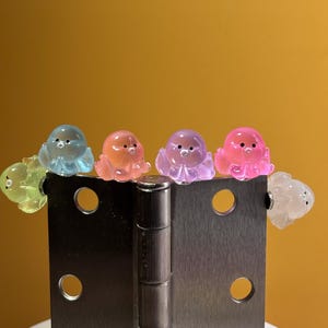 May include: A collection of six small, translucent, octopus-shaped figurines in lime green, light blue, peach, lavender, bright pink, and white, each featuring two tiny black dots for eyes, perched along the top edge of a brushed metal hinge.