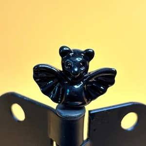 May include: A black bat-shaped decorative element, possibly a cabinet knob or hinge detail, with outstretched wings. The bat has a glossy finish and is mounted on a black metal hinge against a yellow background.