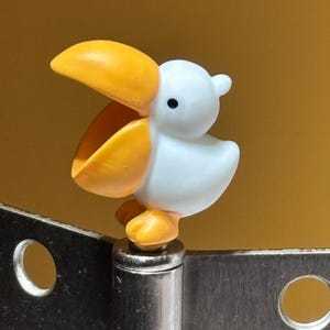 May include: A small, white pelican figurine with a bright orange beak and feet, featuring a wide-open mouth. The bird is mounted on top of a metallic hinge component that includes two circular holes on a flat, rectangular silver-colored plate.