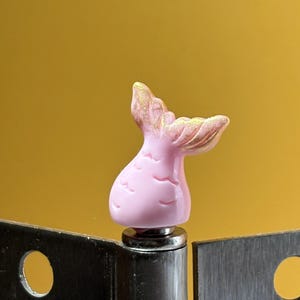May include: A light pink mermaid tail figurine with gold glitter accents on the fin tips. The tail features embossed scale-like markings and is mounted on a small, circular metallic base, positioned between two dark gray metal plates.
