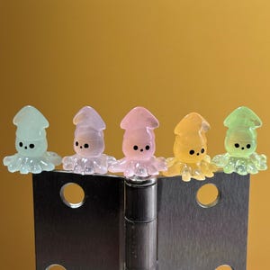 May include: Five small, translucent squid-shaped figurines in light blue, lavender, pink, yellow, and lime green. Each figurine features a smooth, rounded head, a pointed top, and small black dots for eyes, resting atop a metallic hinge.