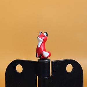 May include: A small, red fox figurine with white accents and black ears sits atop a black hinge. The fox is posed upright, with its tail curled. The background is a solid orange color.