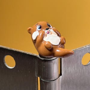 May include: A small, glossy brown and cream-colored otter figurine perched atop a silver metal door hinge. The otter has large, expressive eyes, a small black nose, and is depicted in a reclining position with its paws near its face.