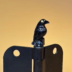 May include: A black crow figurine with yellow eyes sits atop a black metal hinge. The hinge has two rectangular plates with circular cutouts. The background is a solid, warm yellow.