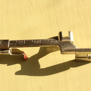 May include: A gold-coloured sewing machine foot with a ruler marked 1, 2, and 3. The foot has a red plastic piece on the side.
