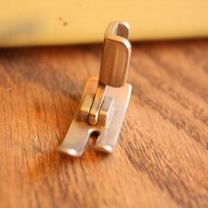 Pode incluir: Um pé de máquina de costura prateado com uma peça de metal elevada na parte superior.