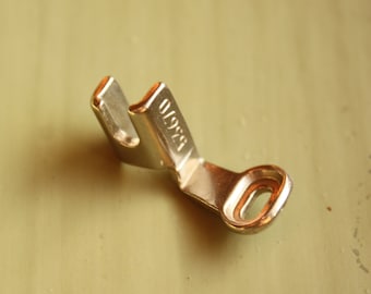 May include: A close-up of a shiny, metallic sewing machine foot. The foot is a light gold color and has a curved shape with a small hole at the end. The number "83670" is stamped on the side.