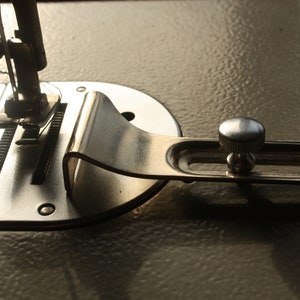 Könnte beinhalten: Nahaufnahme eines silbernen Nähmaschinenfußes mit einem verstellbaren Metallhebel und einem Knopf. Der Fuß ist an einer Nähmaschine mit einem silbernen Metallgehäuse befestigt.
