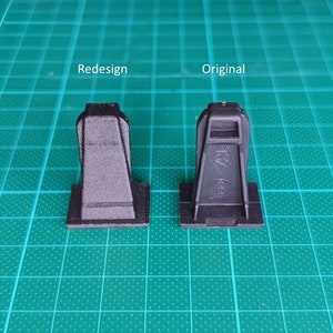 Könnte beinhalten: Zwei schwarze Plastikteile, eines mit der Aufschrift "Original" und das andere mit der Aufschrift "Redesign", werden auf einer grünen Gitteroberfläche gezeigt. Die Teile sind in ihrer Form ähnlich, aber das Teil "Redesign" hat eine abgerundete Oberseite.