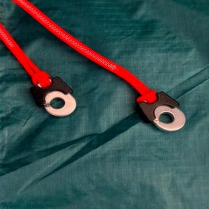 Puede incluir: Dos tiradores de cordón rojo brillante unidos a ojales de metal plateado con conectores de plástico negro, sobre una superficie de tela verde oscuro texturizada tipo ripstop con costuras blancas visibles en el lado izquierdo.
